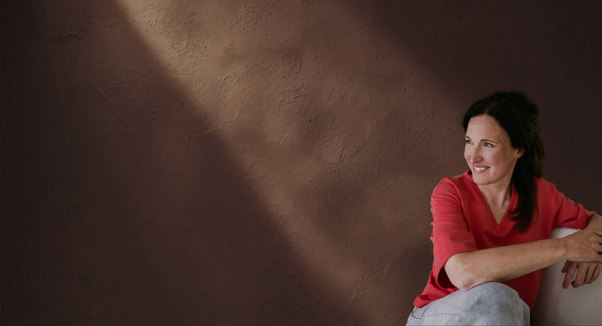 Glimlachende Margo Vermeiren in rood shirt tegen een bruine muur